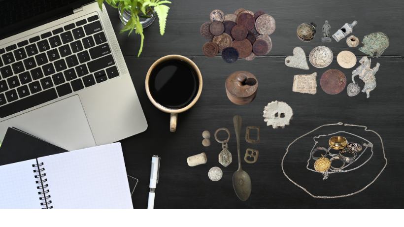 Ausgebreitete Metalldetektor-Funde wie Münzen, Schmuck und Artefakte liegen auf einem Tisch neben Laptop, Notizbuch und Stift – symbolisch für das Dokumentieren und Analysieren von Funden.