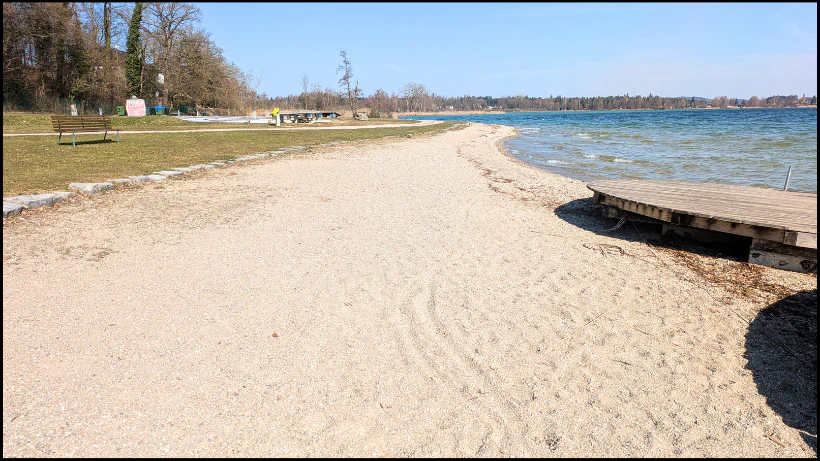 Eine ruhige Liegewiese mit Sandstrand an einem klaren See, daneben ein kleiner Steg und eine gepflegte Grünfläche mit Bänken und Bäumen; typisches neuzeitliches Suchgebiet, an dem viele Menschen Freizeit verbringen und daher moderne Funde möglich sind.