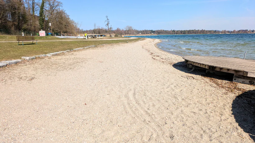 Eine ruhige Liegewiese mit Sandstrand an einem klaren See, daneben ein kleiner Steg und eine gepflegte Grünfläche mit Bänken und Bäumen; typisches neuzeitliches Suchgebiet, an dem viele Menschen Freizeit verbringen und daher moderne Funde möglich sind.