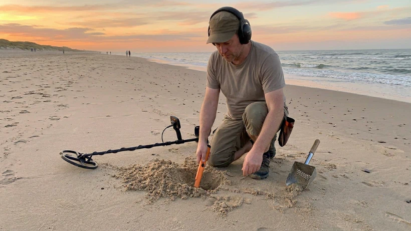 Person sondelt am Strand bei Sonnenuntergang; Metalldetektor, Pinpointer und Sandscoop im Einsatz, kleine Grabung im Sand sichtbar.