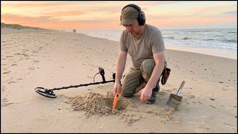 Person sondelt am Strand bei Sonnenuntergang; Metalldetektor, Pinpointer und Sandscoop im Einsatz, kleine Grabung im Sand sichtbar.