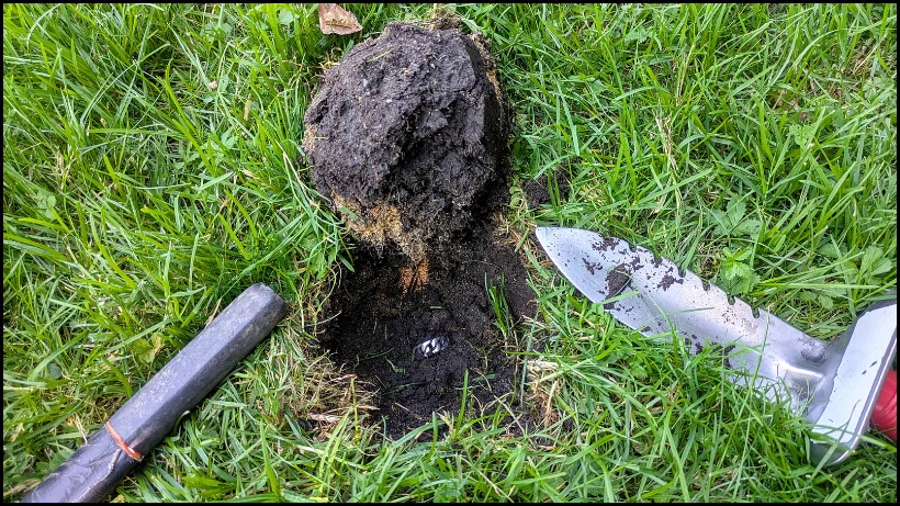 Sauber ausgestochener Plug auf Wiese, Metallfund im Loch sichtbar, Grabungsmesser und Pinpointer zeigen vorsichtiges Graben und sicheres Bergen beim Sondeln.