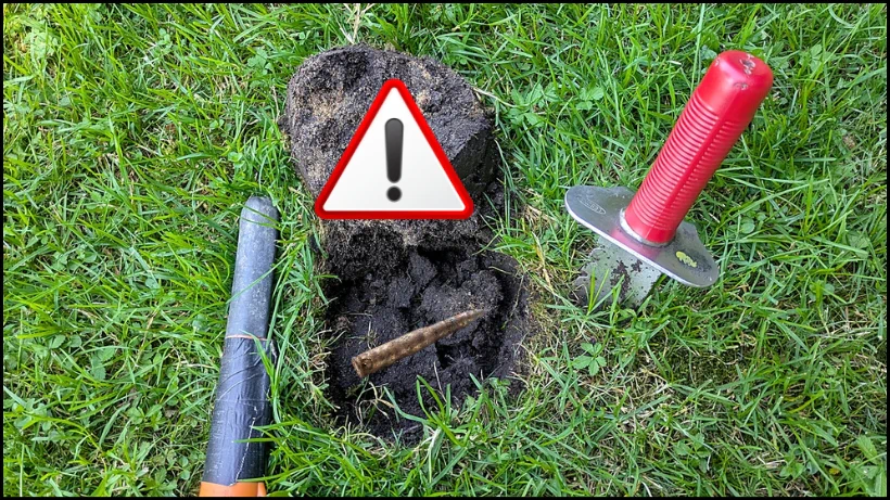 Rostige Gewehrkugel im geöffneten Erdloch, Warnsymbol markiert den Fund, Pinpointer und Grabungsmesser liegen daneben und zeigen vorsichtiges Vorgehen bei gefährlicher Munition beim Sondeln.