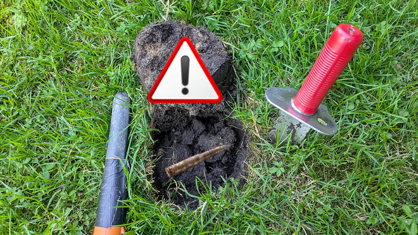 Rostige Gewehrkugel im geöffneten Erdloch, Warnsymbol markiert den Fund, Pinpointer und Grabungsmesser liegen daneben und zeigen vorsichtiges Vorgehen bei gefährlicher Munition beim Sondeln.