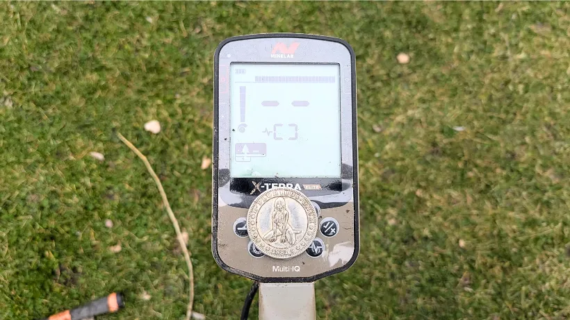 Echter Sondelfund-Beleg: Die detaillierte Hobo-Nickelsilbermünze, graviert mit einer Figur, liegt direkt auf der Multi-IQ Bedieneinheit des Minelab X-Terra Elite Metalldetektors, mit dem sie gefunden wurde. Aufnahme auf Gras-Hintergrund.