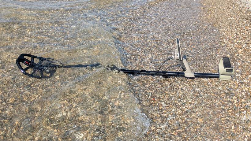 Ein Minelab X-Terra Elite Metalldetektor liegt flach an einem Kieselstrand im flachen Wasser, während eine kleine Welle die V12X-Suchspule und das Gestänge überspült.