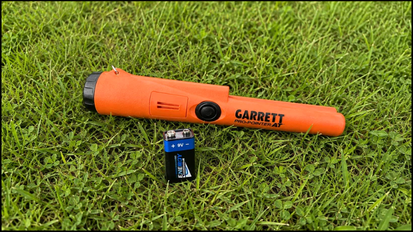 Pinpointer liegt im Gras neben einer 9‑Volt‑Batterie – zeigt Energieversorgung und einfachen Batteriewechsel im Outdoor‑Einsatz.