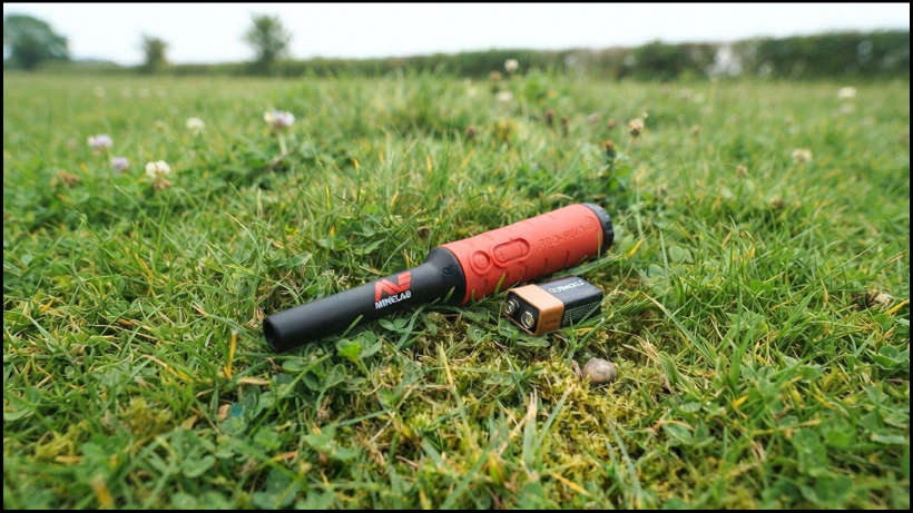 Minelab Pro‑Find 35 liegt im Gras neben einer 9V‑Batterie, als Hinweis auf die Stromversorgung und typische Batterielaufzeit im Einsatz.