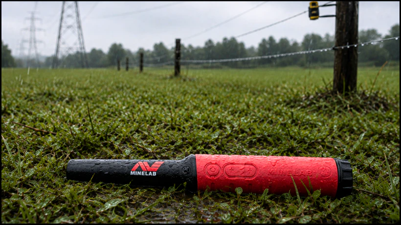 Minelab Pro‑Find 40 liegt im nassen Gras nahe Stromzäunen und Leitungen, um Stabilität und mögliche Störanfälligkeit durch EMI im Außeneinsatz zu zeigen.