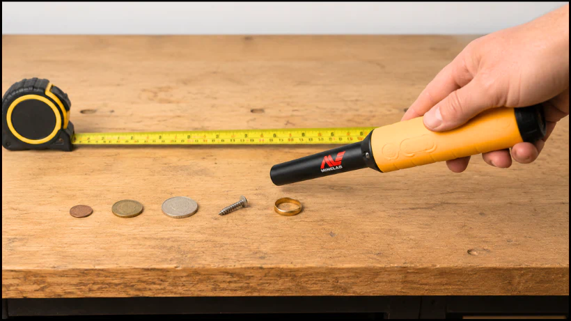 Pro‑Find 35 Pinpointer im Indoor‑Testaufbau auf Holztisch, daneben ausgerolltes Maßband, verschiedene Münzen, Schraube und Goldring, realistische Darstellung der Messergebnisse zuhause unter kontrollierten Bedingungen.