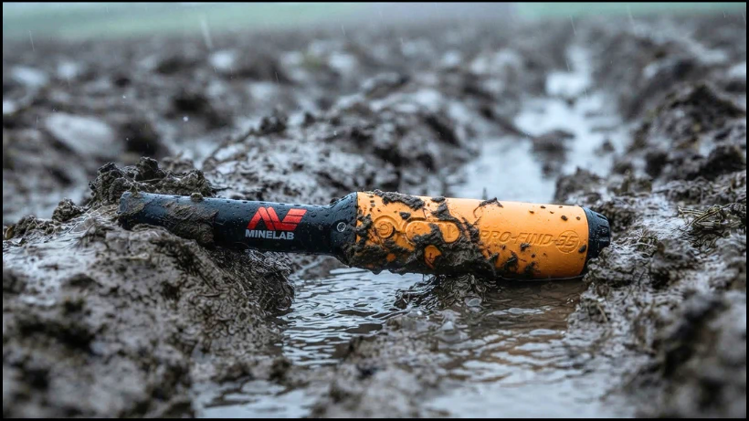Minelab Pro‑Find 35 liegt verschmutzt im nassen Erdloch, Darstellung der robusten Bauweise und vollständigen Wasserdichtigkeit bei Regen, Schlamm und feuchten Böden.